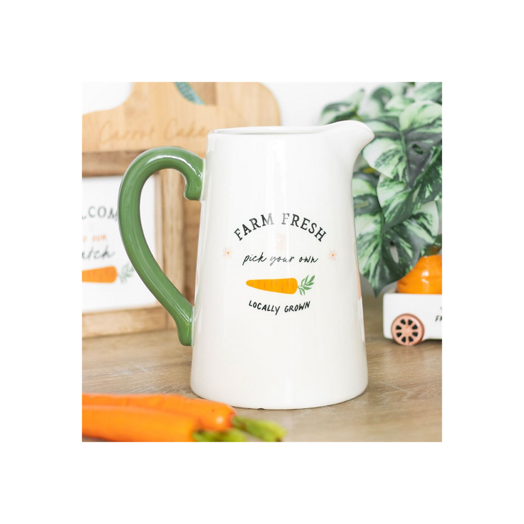 White pitcher with green handle on a wooden surface with carrots and plants in the background