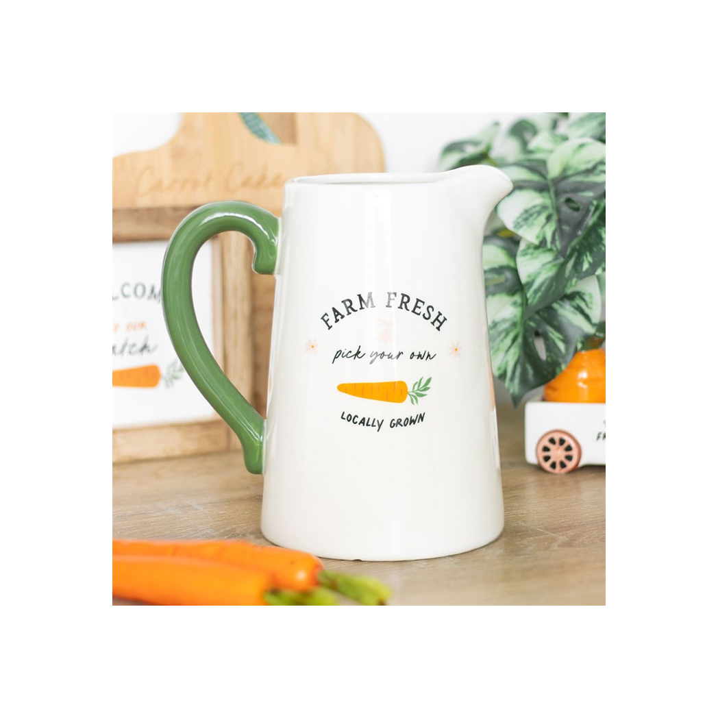 White pitcher with green handle on a wooden surface with carrots and plants in the background