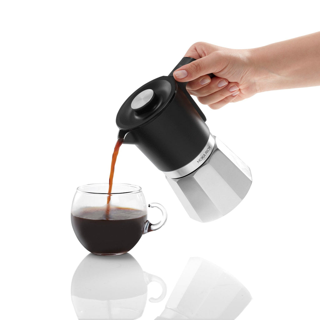 Hand pouring coffee from a black and silver coffee maker into a glass mug on a white background