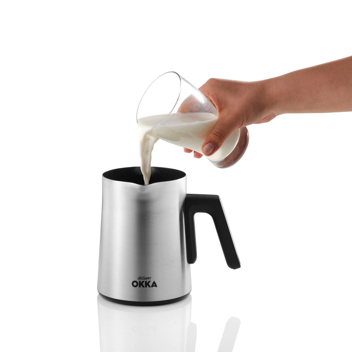 Person pouring milk from a glass into a stainless steel milk frother with black handle on a white background.