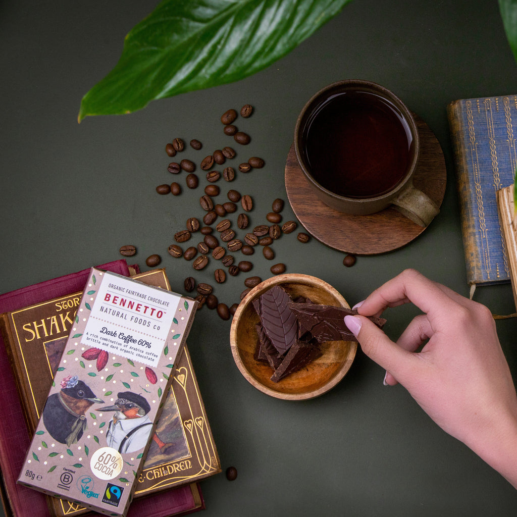 Hand holding a chocolate bar over a cup of coffee with coffee beans and books on a dark surface.