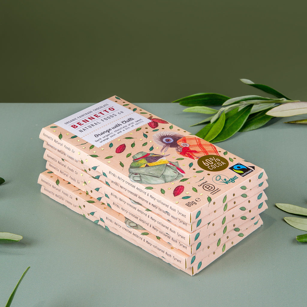 A stack of Bennetto chocolate bars orange-flavored, wrapped in pale pink paper with green leaf patterns and illustrations, displayed on a light sage green surface with olive branches in the background.
