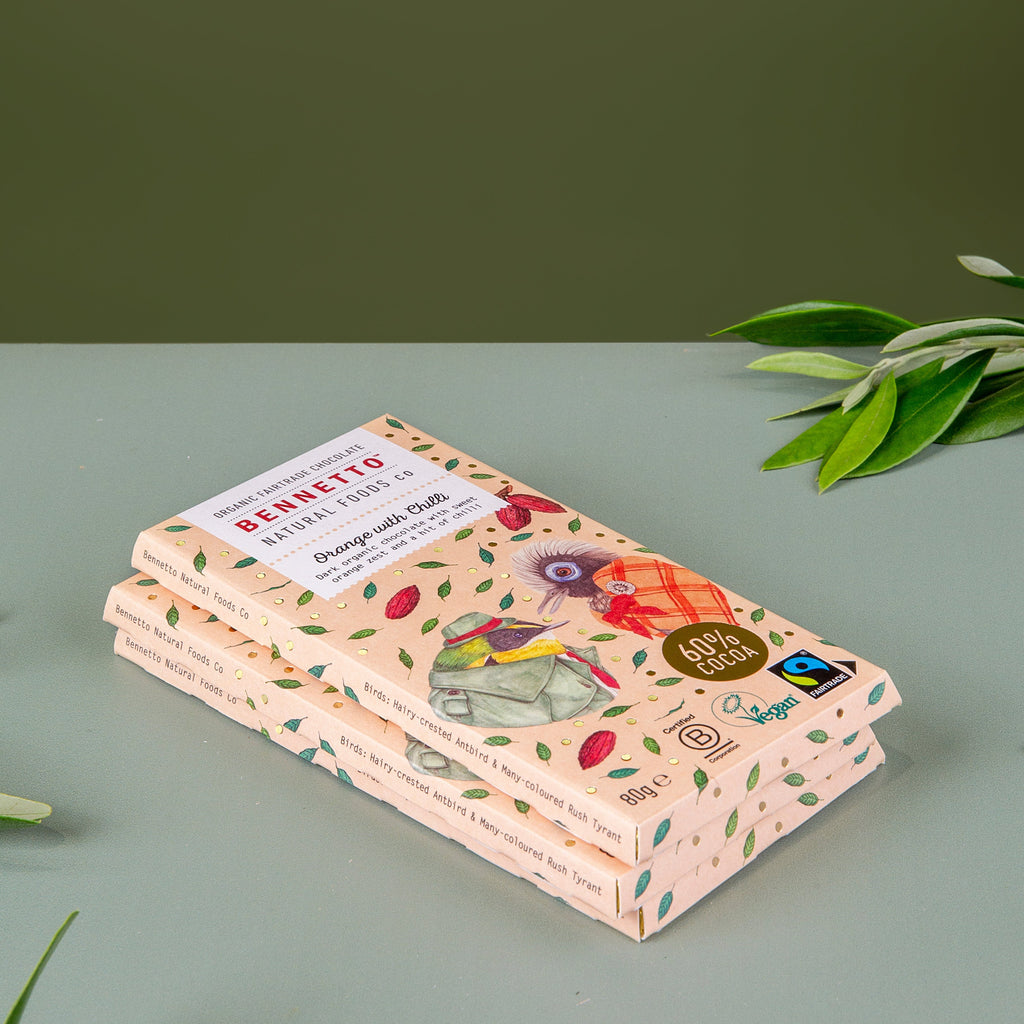 Three stacked bars of Bennetto Organic Cocoa Chocolate in a light peach packaging with green leaf patterns and colourful illustrations, set on a gray surface with a dark green background and olive branches.