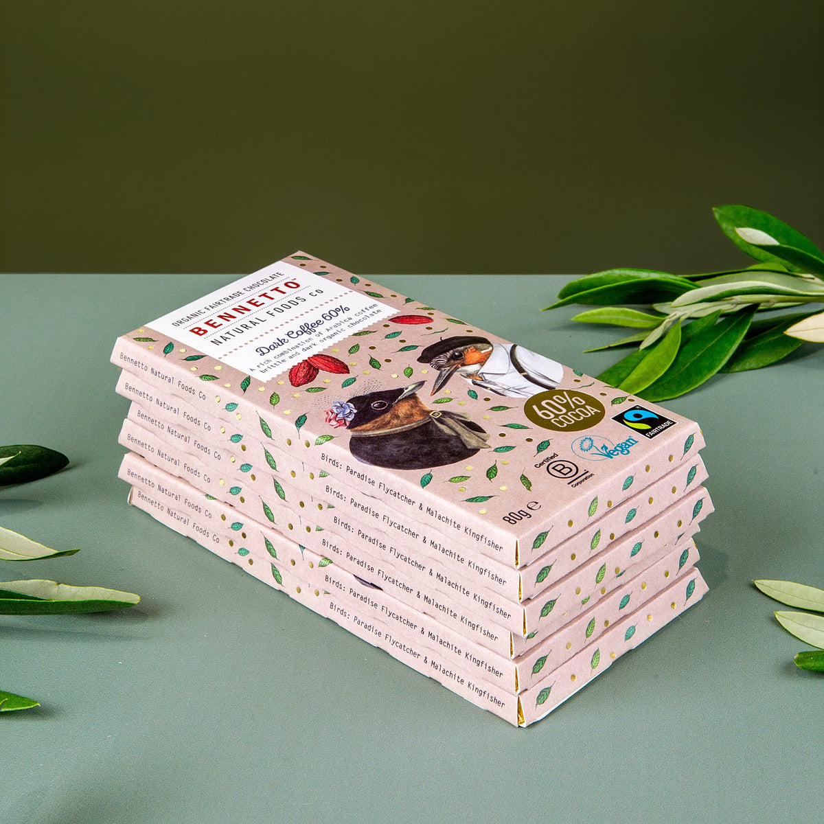 A stack of Genato chocolate bars with a light pink wrapper decorated with green leaves and illustrations, styled on a sage green surface with olive branches.