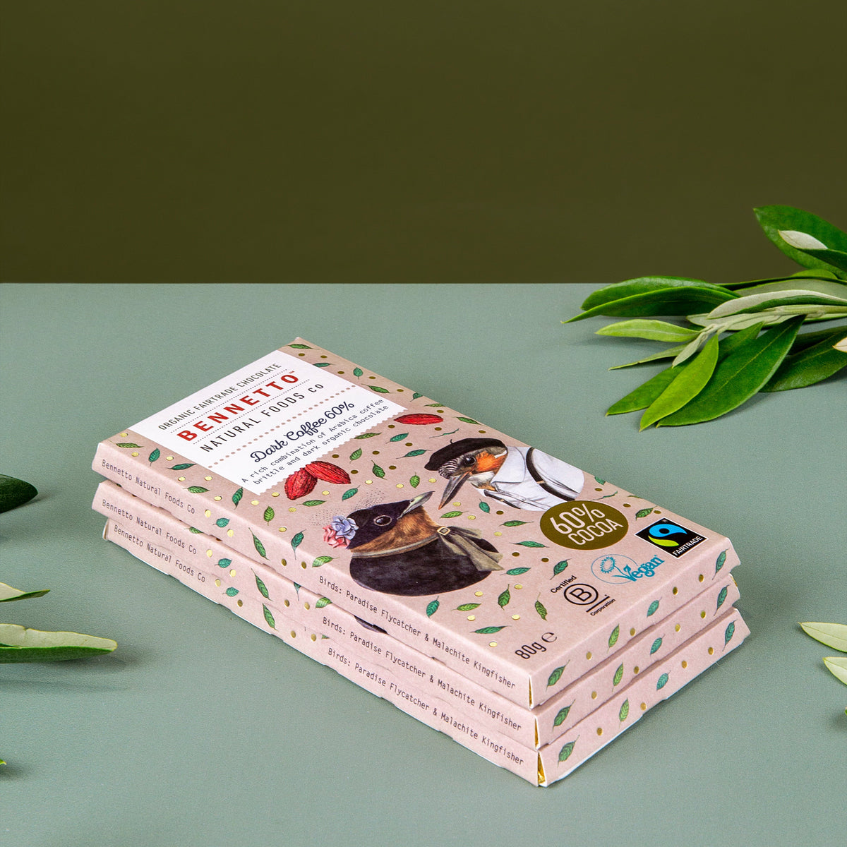A stack of four Berretto chocolate bars in light pink packaging with green leaf patterns, styled on a sage green surface with olive leaves against a dark olive background.