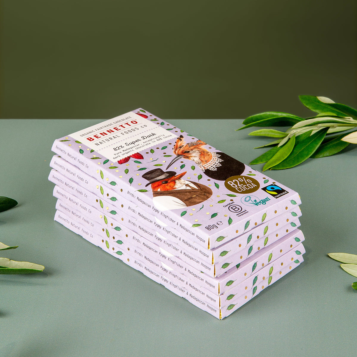 A stack of Benetton chocolate bars in light pink packaging with colorful illustrations, styled on a muted green surface with green leaves.