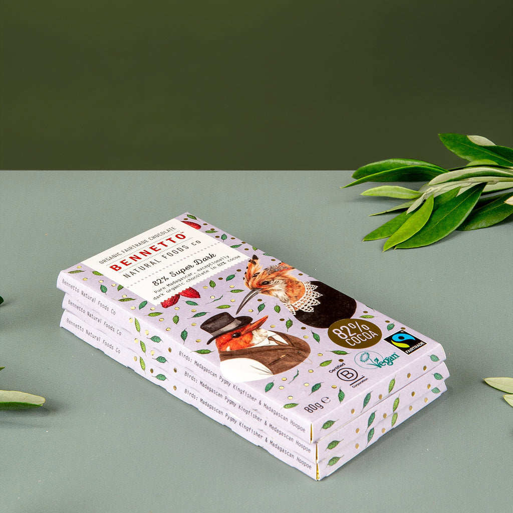 A stack of chocolate bars in decorative white wrappers with green leaf patterns and illustrations, styled on a light gray surface with olive leaves against a dark olive green background.