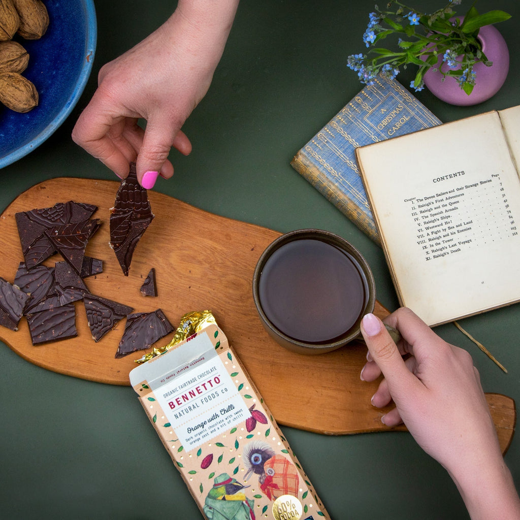 Person holding a piece of chocolate over a cup of tea with a Bennett's chocolate bar on a wooden board.