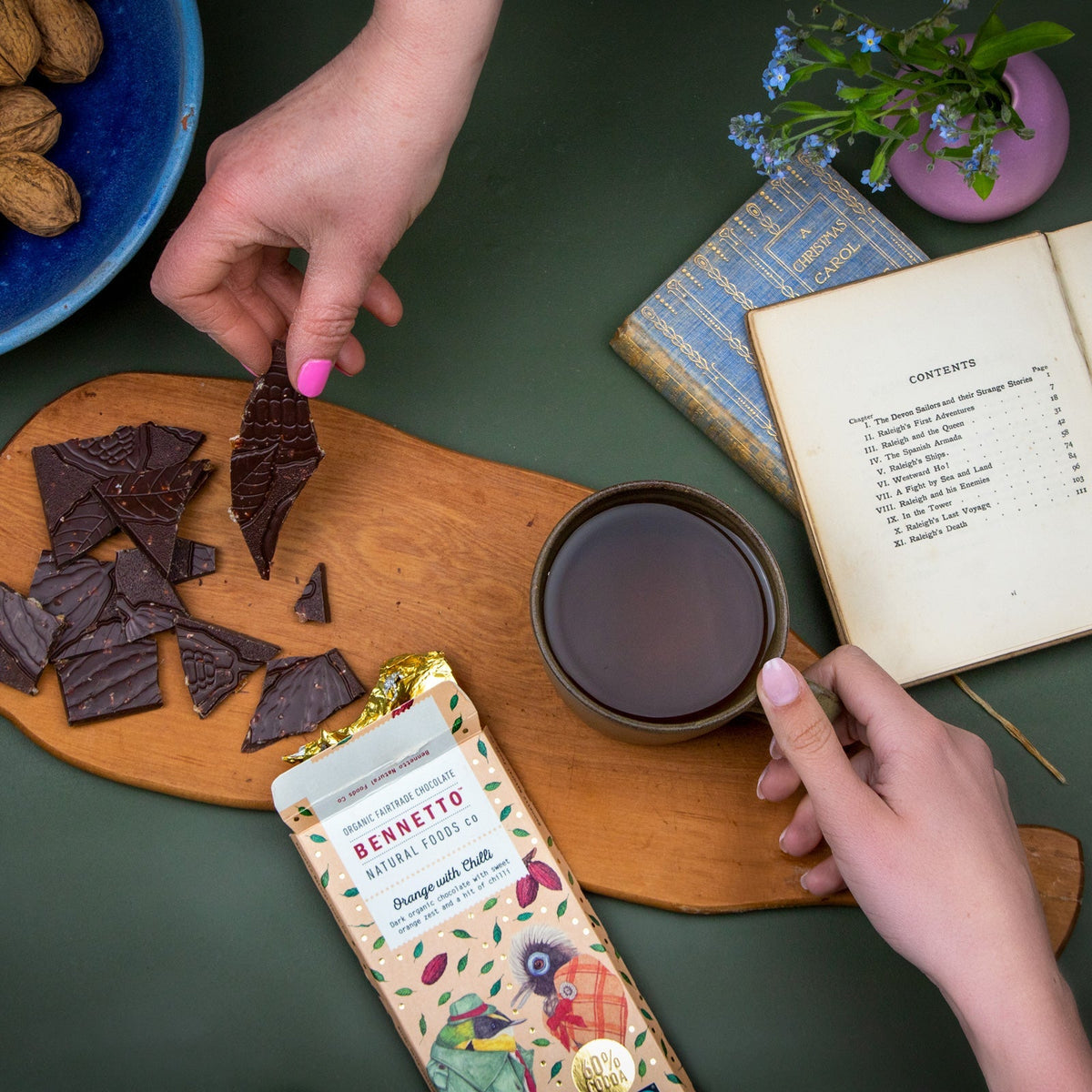 Person holding a piece of chocolate over a cup of tea with a Bennett's chocolate bar on a wooden board.
