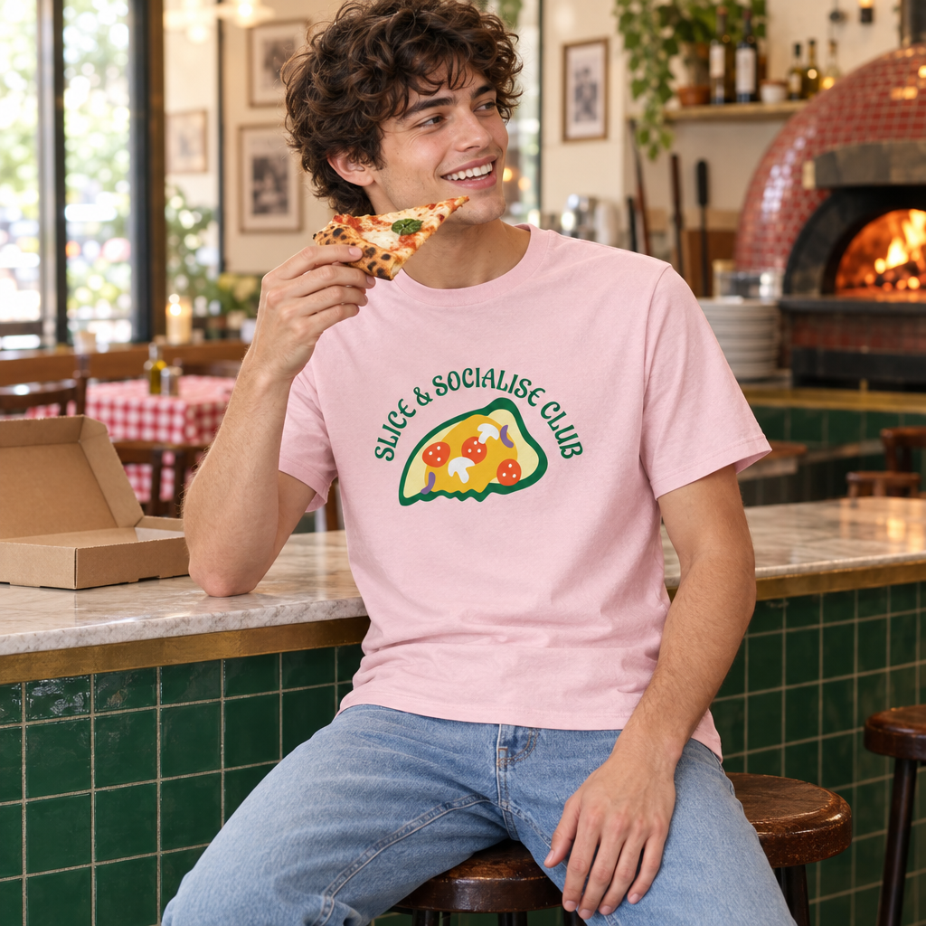 A smiling man in a pink graphic t-shirt and blue jeans sits at a counter in a pizza shop, holding a slice of pizza. The t-shirt features a green graphic of a slice of pizza with the text 'SLICE & SOCIALISE CLUB'.
