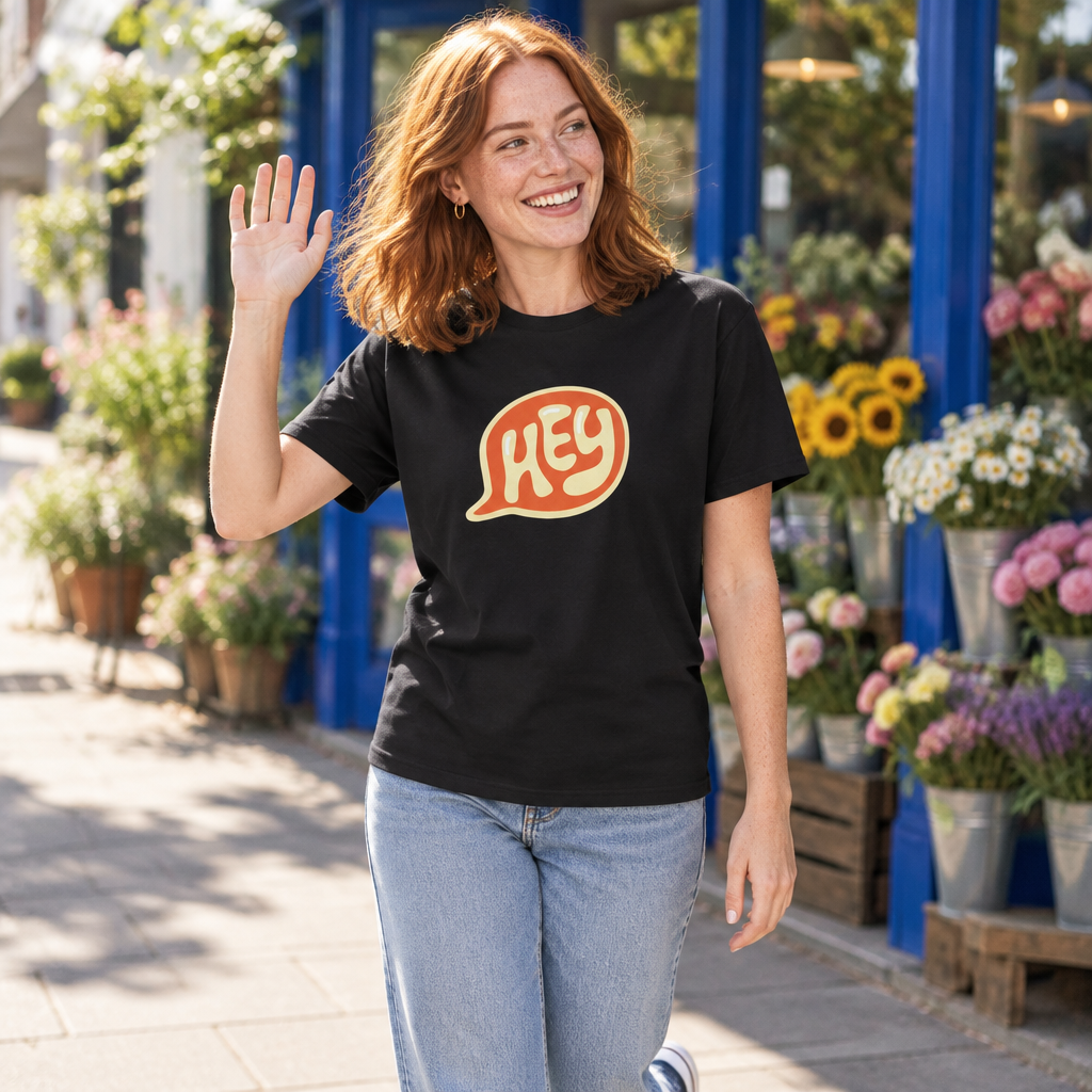 A woman with red hair smiling and waving while wearing a black t-shirt with a retro speech bubble graphic that says 'HEY' in orange and yellow. She is standing outdoors in front of a flower shop with blue frames and potted flowers.