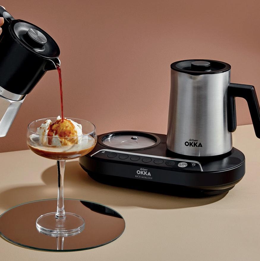 Black and silver OKKA coffee maker with a glass of dessert being poured over it on a beige background