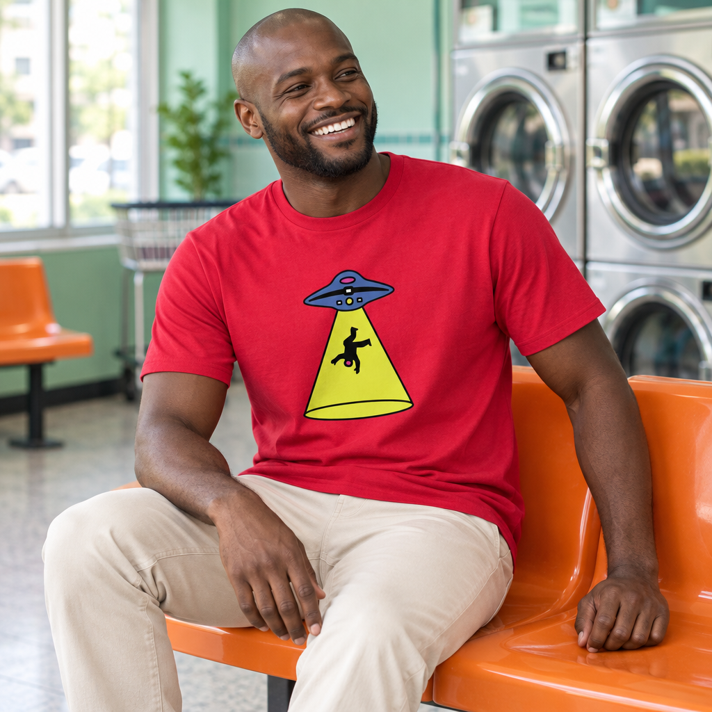 A smiling man wearing a bright red graphic t-shirt sits on an orange bench in a laundromat. The t-shirt features a cartoon graphic of a UFO with a yellow spotlight shining down.