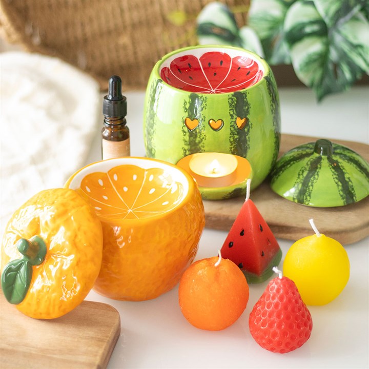 Fruit-themed candles including watermelon, orange, and strawberry on a wooden surface with a plant in the background.