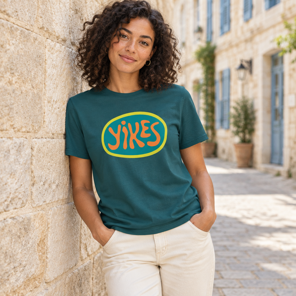 A woman with curly hair stands against a stone wall wearing a dark teal graphic t-shirt with an orange and yellow 'YIKES' logo, paired with cream-colored pants in an outdoor street setting.