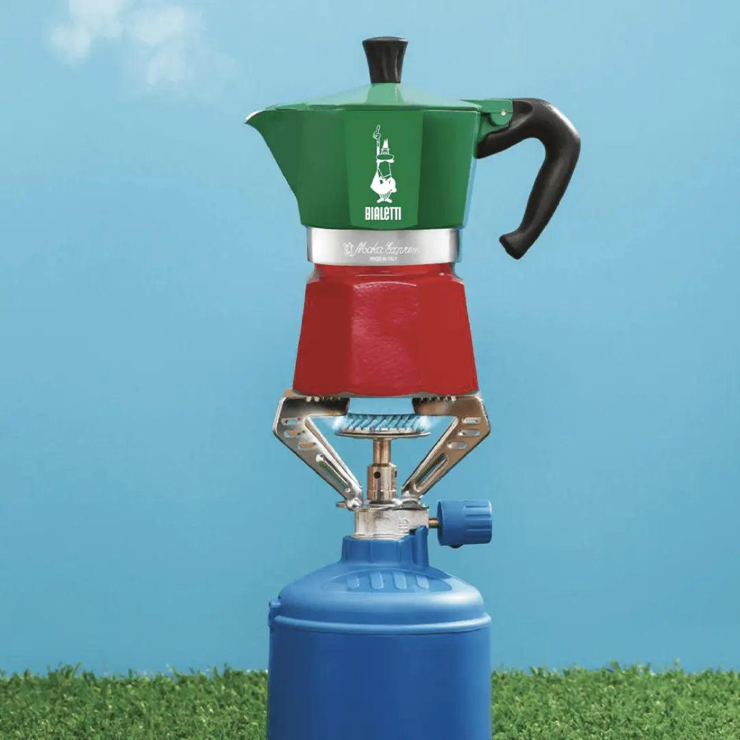 Green and red coffee maker on a blue camping stove against a blue sky background