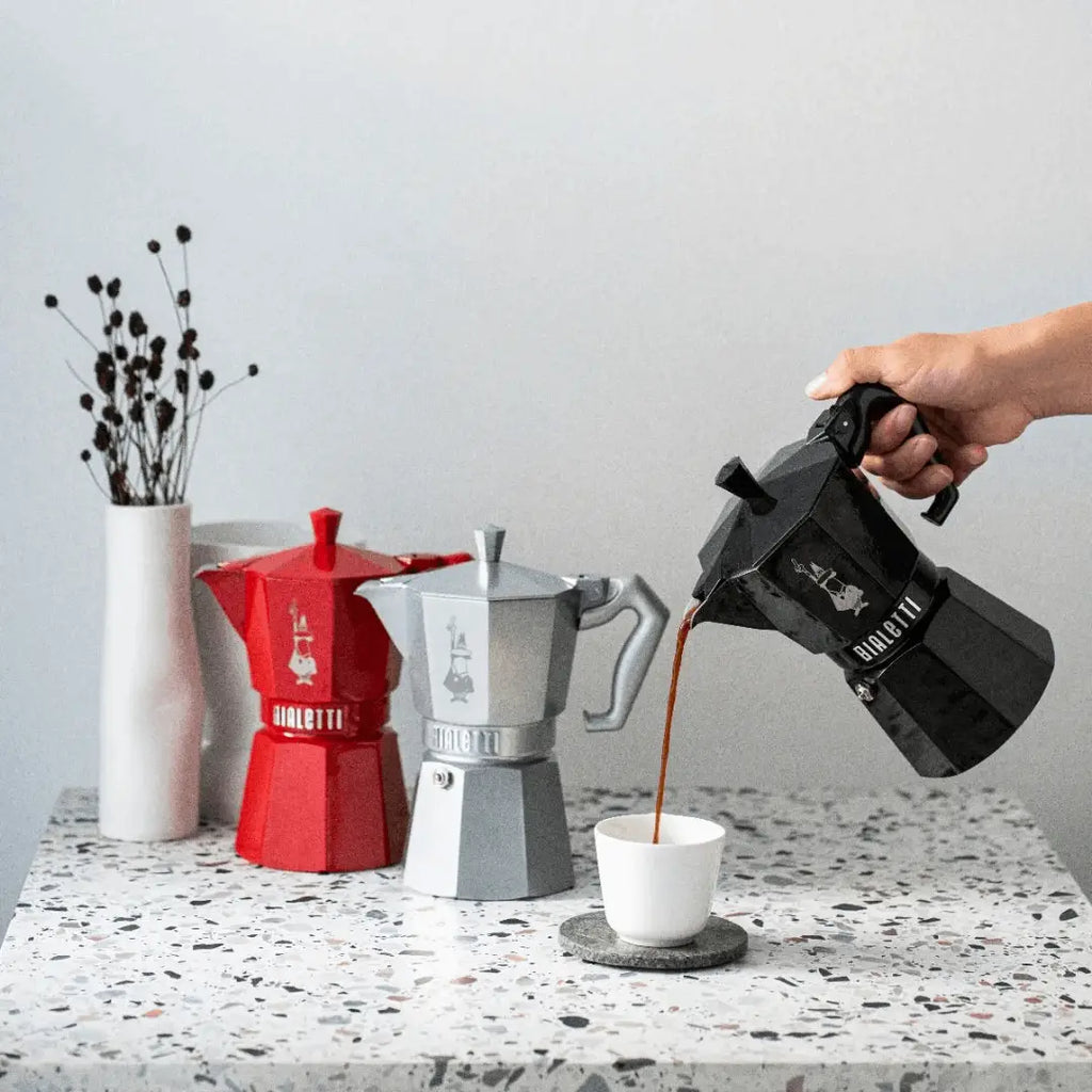Person pouring coffee from a black Moka pot into a white cup on a speckled counter top with red and silver Moka pots in the background.
