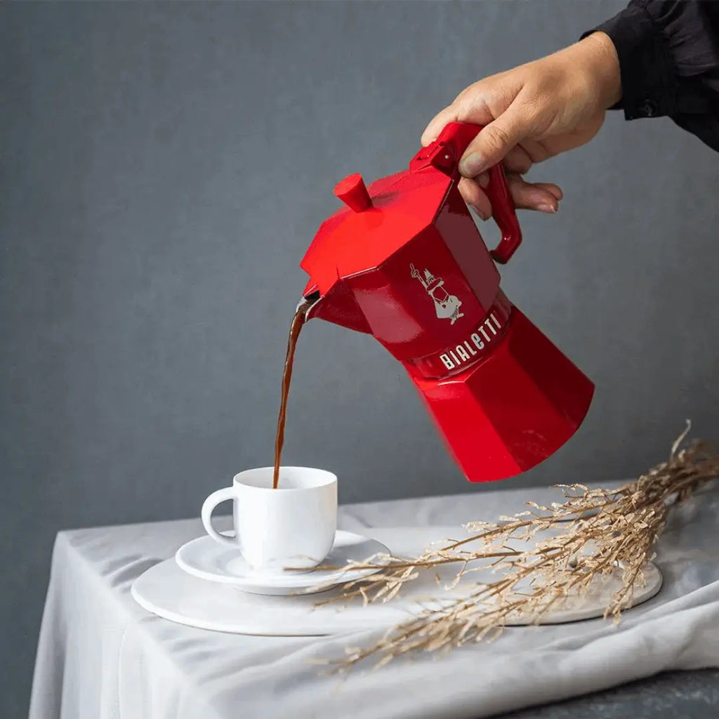 Red Bialetti coffee maker pouring coffee into a white cup on a gray background
